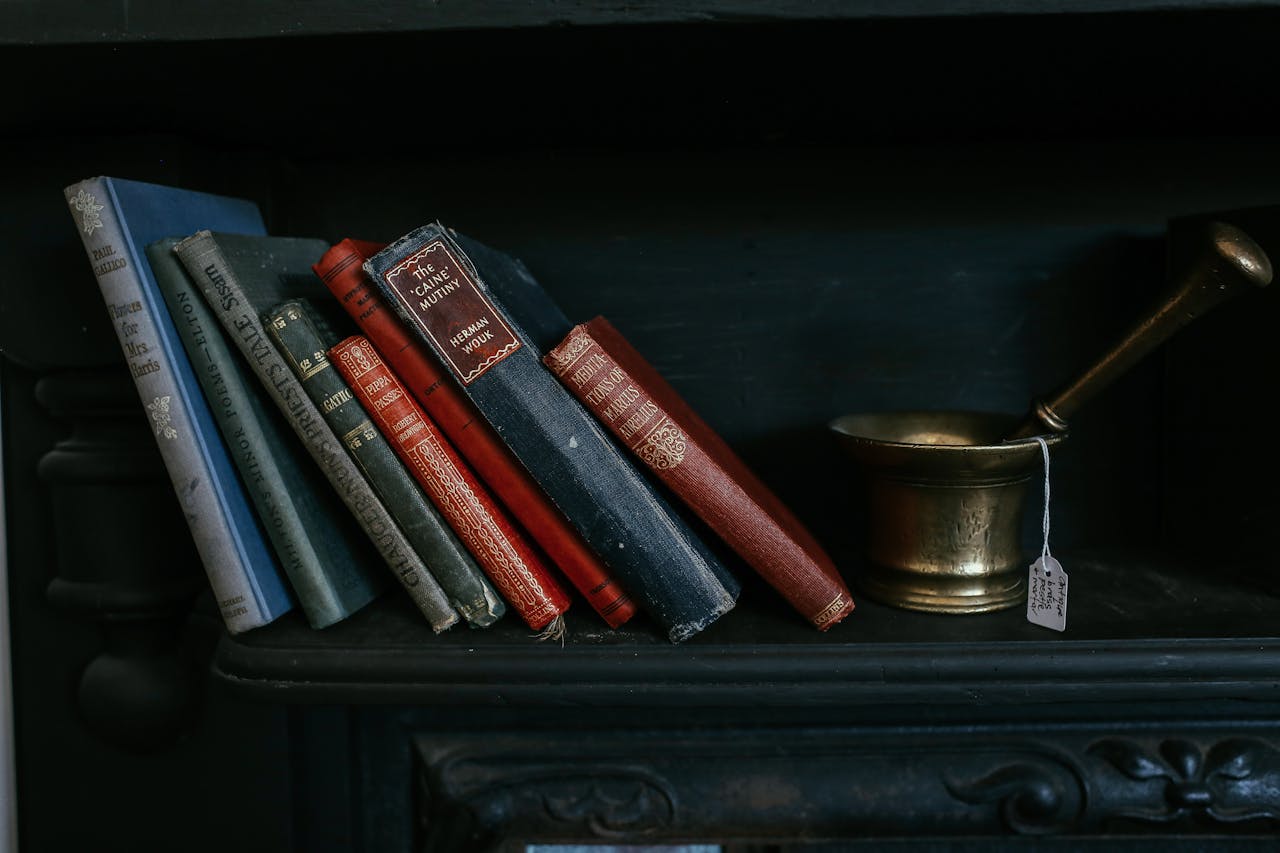 A collection of vintage books and a brass mortar on a dark shelf creates a classic still life scene.